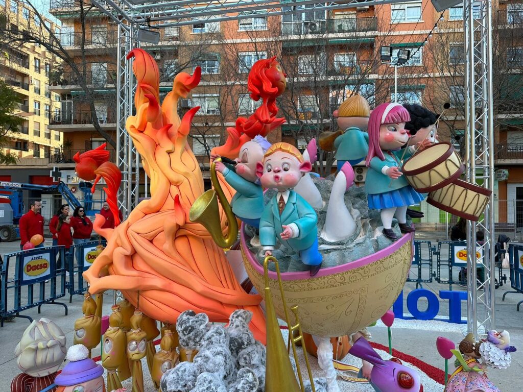 Falla infantil. Hay ninots de falla que son niños y niñas que forman parte de una banda de música. Los niños y las niñas tocan el tambor, el trombón y el saxofón. Todos los niños y niñas de la banda están subidos a una barca. Y detrás tienen una llama de fuego. Al fondo de la imagen hay gente viendo la falla.