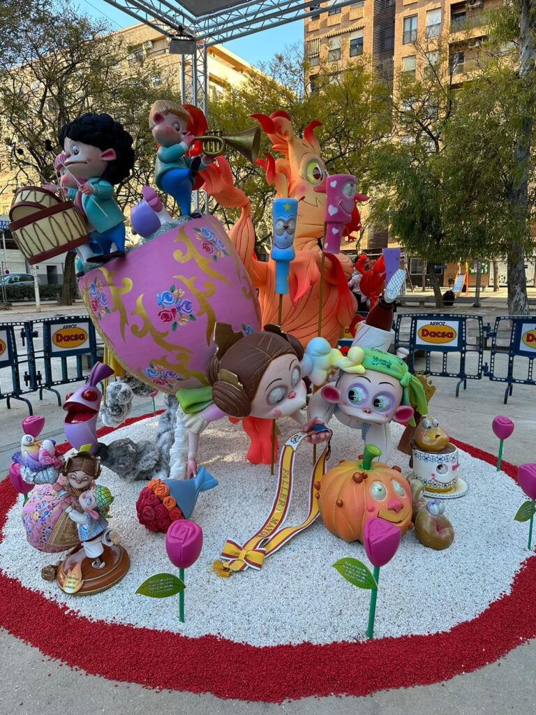 Falla infantil. En la imagen principalmente se ven un niño fallero y una niña fallera, tumbados y mirando a una calabaza con ojos, nariz y boca. Encima de la niña fallera hay unos niños tocando el tambor y la trompeta. Al fondo de la imagen se ve una llama de fuego con ojos, nariz y boca. También se ven unas rosas de color fucsia, que rodean toda la falla infantil.