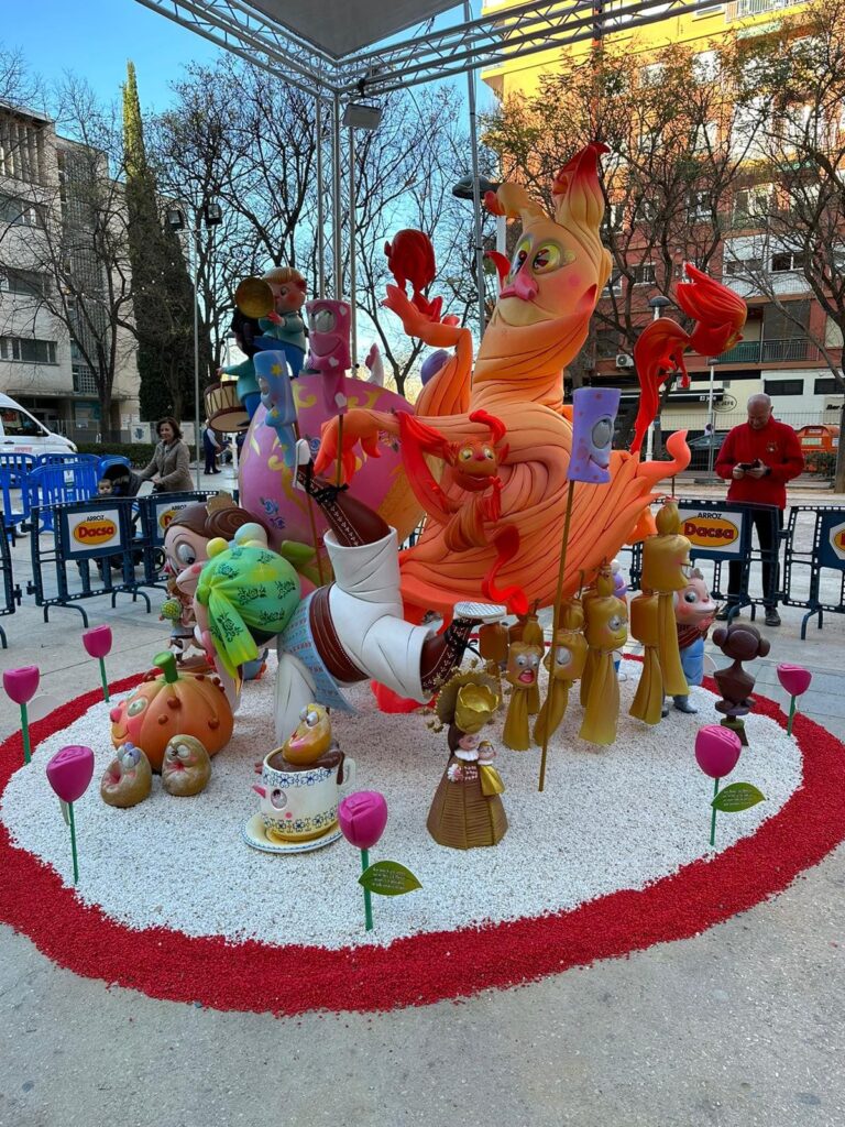 Falla infantil. El ninot principal es una llama de fuego con ojos, nariz y boca. También hay unos niños falleros mirando a una calabaza con ojos, nariz y boca, y al lado una serie de masclets con ojos, nariz y boca, además de una taza pequeña y una figura se la Mare de Deu de Valéncia. También se ven unas rosas de color fucsia, que rodean toda la falla infantil.