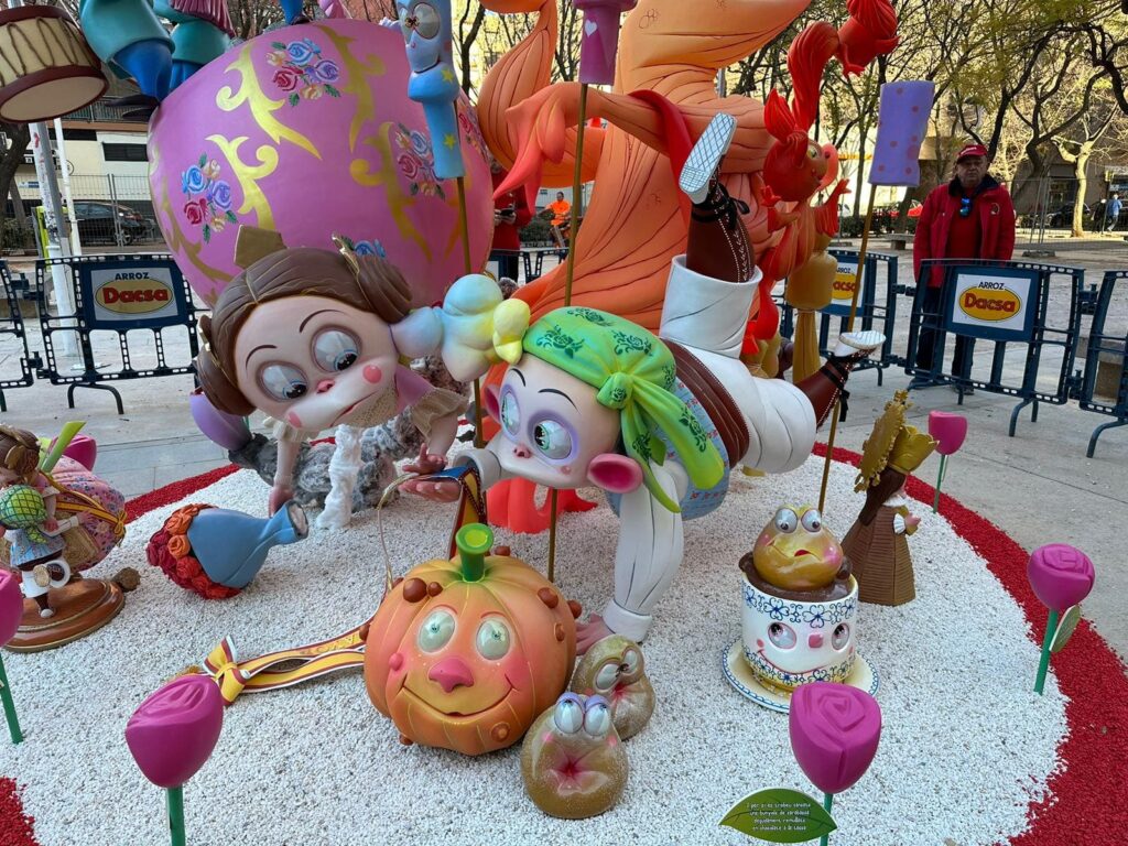 Falla infantil. Son un niño fallero y una niña fallera que están tumbados mirando a una calabaza que tiene ojos, nariz y boca. Delante y en el suelo, hay una taza pequeña, y una figura de la Mare de Deu de Valéncia. Y Además unas rosas de color fucsia, que rodean toda la falla infantil.