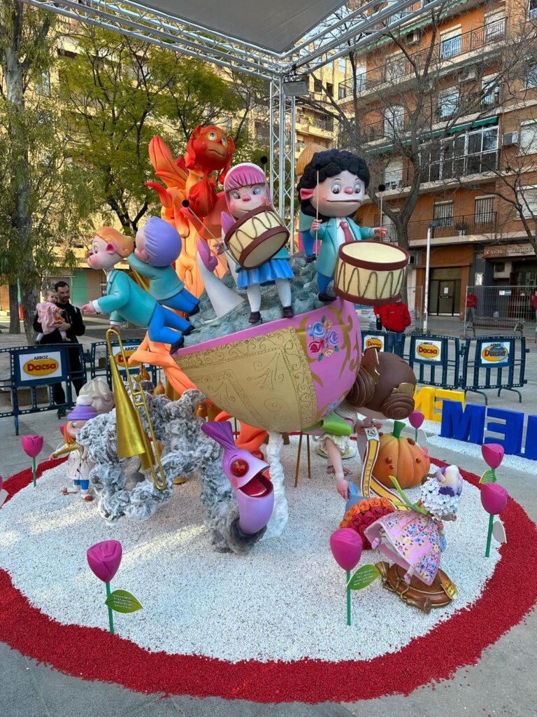 Falla infantil. En la imagen principalmente se ven una niña y un niño tocando el tambor encima de una barca. Delante y en el suelo, hay un masclet y unas rosas de color fucsia, que rodean toda la falla infantil.