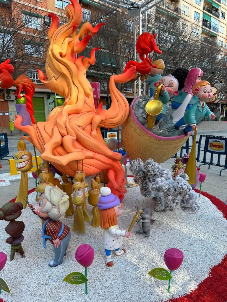 Falla infantil. El ninot de falla principal es una llama de fuego gigante. Delante se ven unos ninots pequeños en el suelo, de un pintor y un escultor. Detrás de la llama de fuego, y al fondo de la imagen, hay unos niños y niñas que forman parte de una banda de música, que están subidos a una barca.