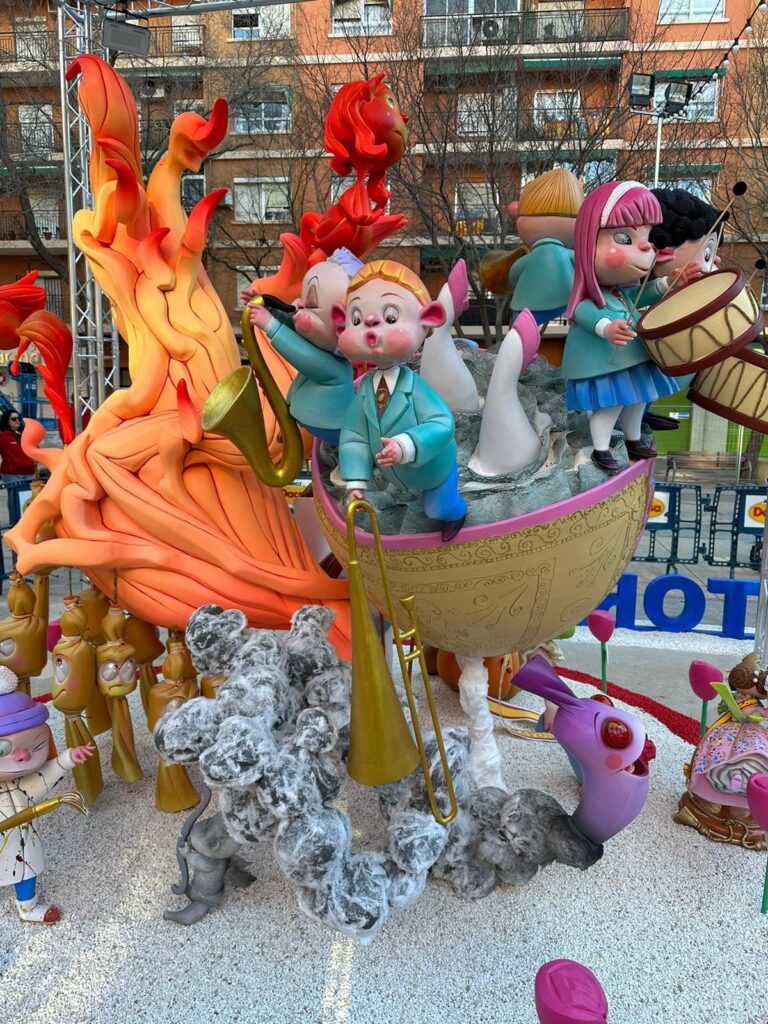 Falla infantil. Hay ninots de falla que son niños y niñas que forman parte de una banda de música. Los niños y las niñas tocan el tambor, el trombón y el saxofón. Todos los niños y niñas de la banda están subidos a una barca. Y detrás tienen una llama de fuego bastante grande.