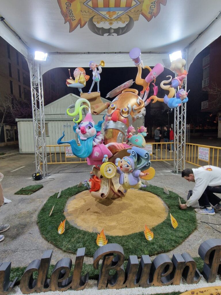 Falla infantil. El ninot principal es un catalejo grande con ojos, nariz y boca, al cual le rodean los otros ninots: un payaso con barza azul y traje rosa, un niño con un reloj despertador, y encima una niña tocando el tambor, y un niño tocando la trompeta. En frente y en el suelo hay un cartel en el que pone: del Buen. Parece que el cartel sigue, pero no se ve en la imagen.