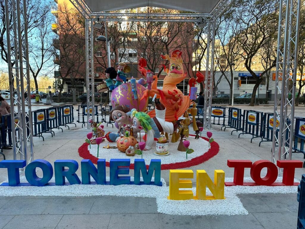 Falla infantil. El ninot principal es una llama de fuego con ojos, nariz y boca. Debajo hay un niño fallero y una niña fallera, que están tumbados mirando a una calabaza que tiene ojos, nariz y boca. Delante de la falla y puesto en el suelo, hay un cartel que pone: TORNEM EN TOT.