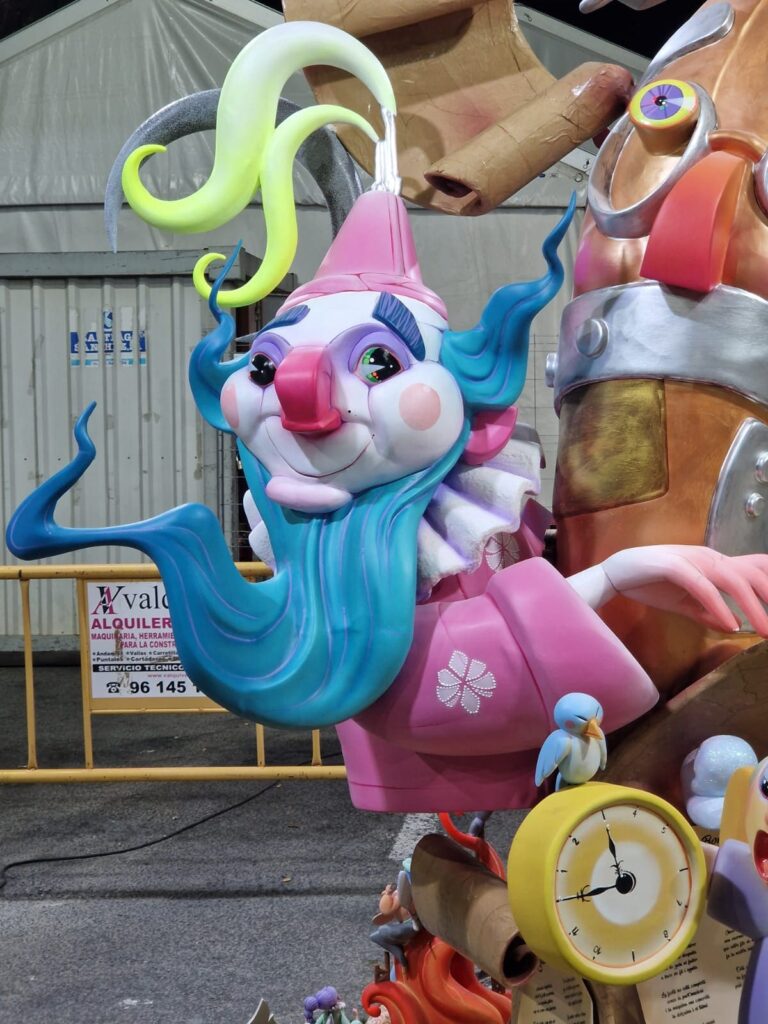 Ninots de falla. Es el ninot de un payaso con barba azul y traje rosa. Delante se ve un reloj despertador y un pájaro azul posado encima del reloj despertador.