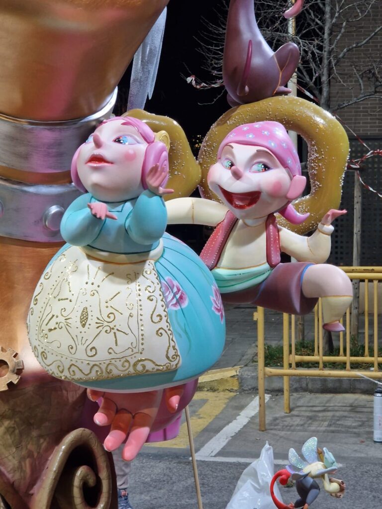 Ninot de falla. Es un niño fallero y una niña fallera que están encima de la mano de otro ninot de falla más grande. La fallera lleva un vestido azul, y el pelo de color rosa.