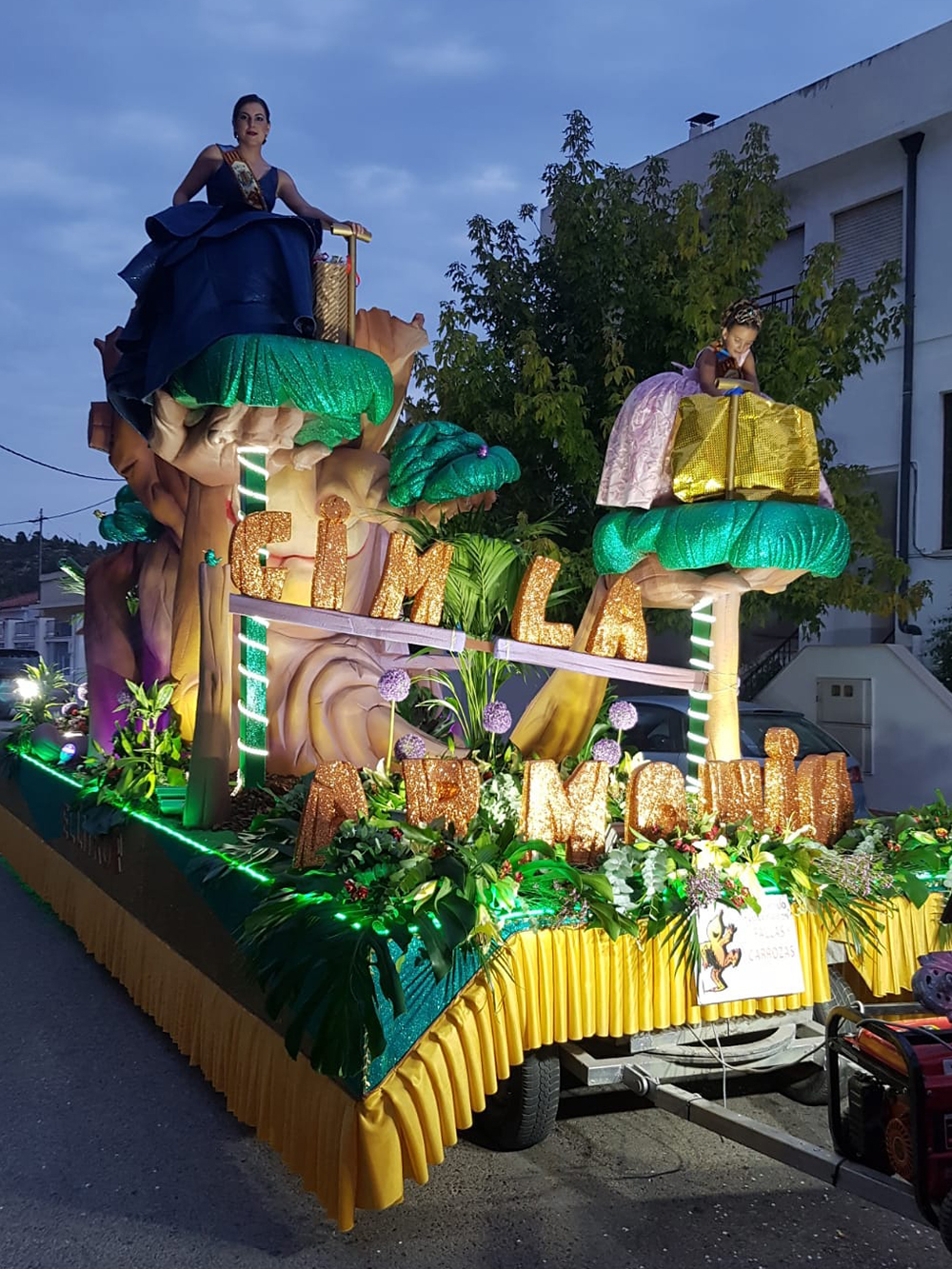 Carroza. Es una carroza decorada con árboles, troncos y plantas pequeñas. Delante de la imagen se ve el letrero que está en el suelo de la carroza que pone: CIM LA ARMONICA. Es de noche, y la carroza está desfilando por la calle. Encima de los árboles más grandes de la carroza están sentadas, una niña y una mujer, que llevan un vestido muy elegante.