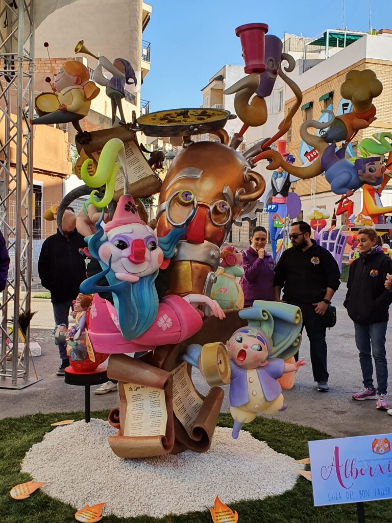 Falla infantil. El ninot principal y más grande de la falla, es un catalejo con ojos, nariz y boca, al que rodean otros ninots de falla. Un payaso con barza azul y traje rosa, un niño con un reloj despertador, y encima del catalejo hay una paella de arroz con pollo y unos niños tocando el tambor y la trompeta. Al fondo de la imagen hay gente mirando la falla.