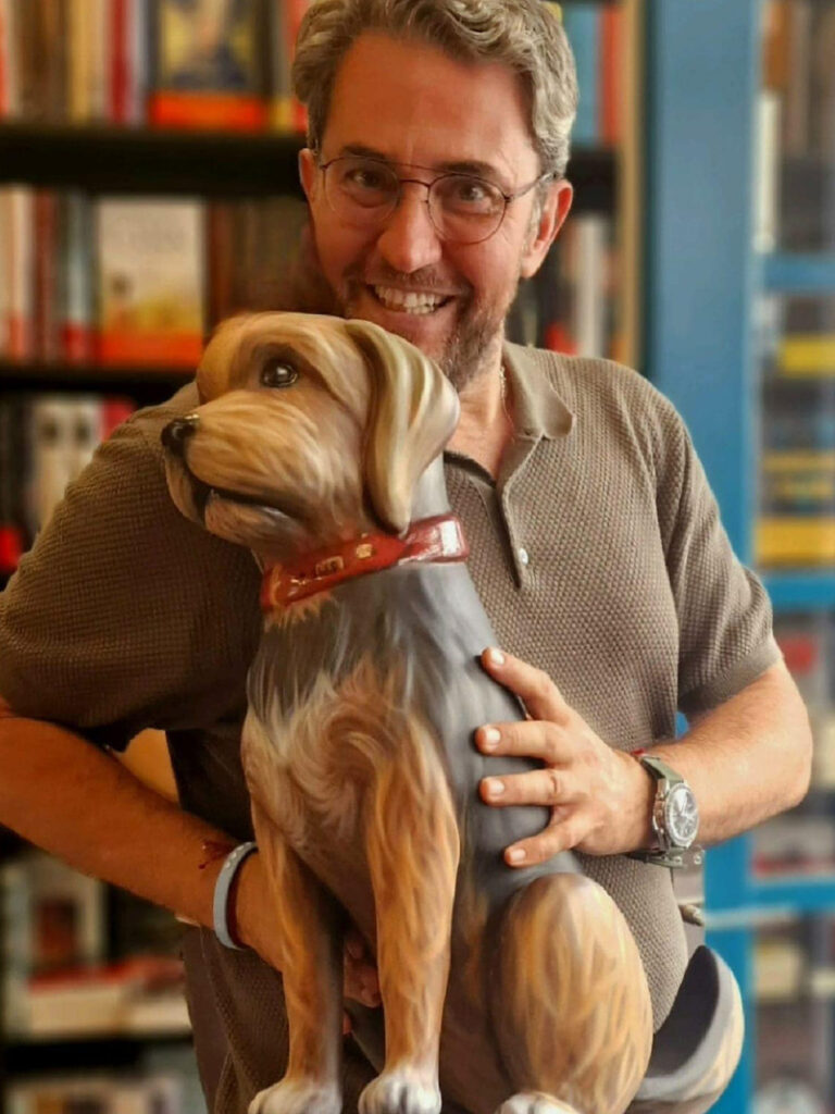 Es Màxim Huerta, y está sonriendo. Sostiene un ninot de falla de un perro, que representa a su perrita y el ninot está hecho a tamaño real.